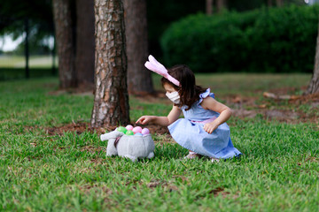 Little toddler girl with bunny ears and surgical face mask hunting for Easter eggs during coronavirus quarantine - 337056845