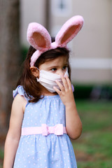 Little toddler girl with bunny ears and surgical face mask hunting for Easter eggs during coronavirus quarantine - 337057033