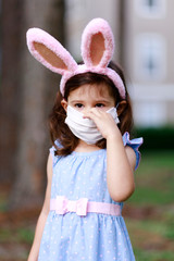 Little toddler girl with bunny ears and surgical face mask hunting for Easter eggs during coronavirus quarantine - 337057071