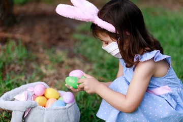 Little toddler girl with bunny ears and surgical face mask hunting for Easter eggs during coronavirus quarantine - 337057276