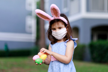 Little toddler girl with bunny ears and surgical face mask hunting for Easter eggs during coronavirus quarantine - 337057288