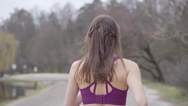 Young healthy Caucasian woman jogging in park on lake shore. Back view of slender brunette girl running along the alley on cloudy spring or autumn day. Sport, lifestyle, athleticism, workout.