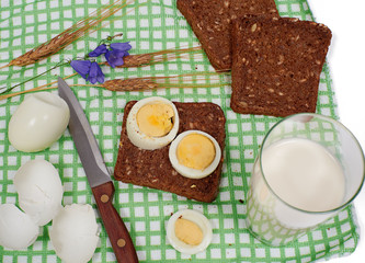 Slices of bread with eggs, glass of milk, rye and bluebell flower on checkered table-napkin