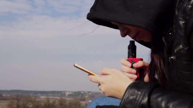 Close up of young woman in hood vaping and flipping through news in mobile phone, sitting on high ground in countryside.