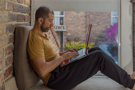 Young Man With Beard, Sweat Pants, T-shirt And His Laptop Over The Legs Working At Home By A Window. Home Office Concept