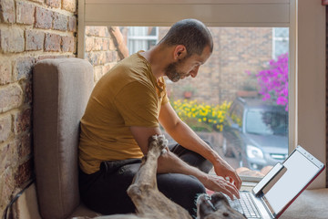 Young man in sweat pants and t-shirt typing in his laptop while his dog ask for attention. Home office concept