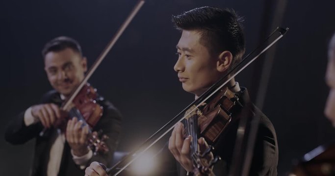 Close up of smiled hapy male young Asian violinist playing on violin in classical orchestra with Caucasian colleague at concert. Cheerful musician performing on his instrument.