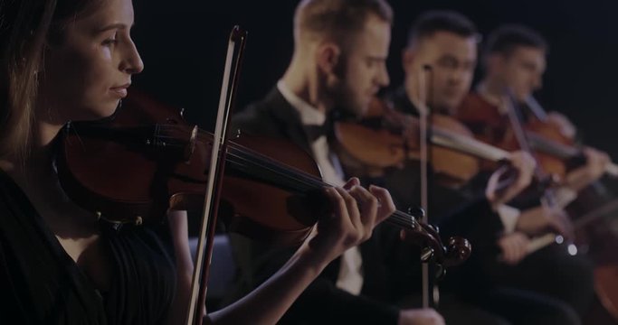 Close up of female young Caucasian violinist playing on violin in classical orchestra with male colleagues at concert. Musician performing on her instrument.