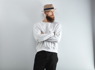 Serious bearded man in hat standing against white wall arms crossed.