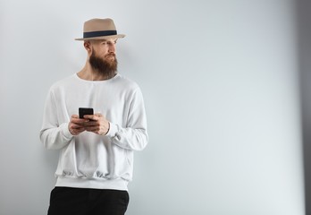 Serious bearded man in hat standing against white wall using smart phone.