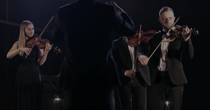 Rear of male elegant conductor controlling band. Quartet of classical male and female musicians performing their music on dark stage in spotlight. Orchestra playing on violins and cello. Back view.
