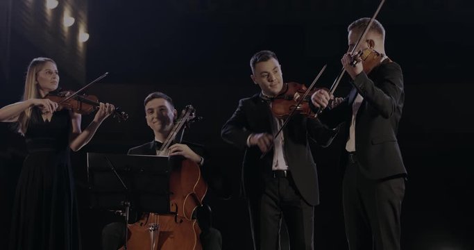 Cheerful men and woman from quartet of classical music having a concert. Male and female musicians performing their music on dark stage in spotlight. Orchestra playing on violins and cello.