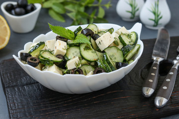 Healthy salad with cucumbers, feta cheese and black olives, with olive oil and greens, located in white bowl on a dark background, horizontal orientation, selective focus