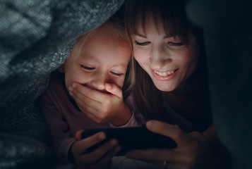 Stay home. Mother with a little daughter watching content on  smartphone in the dark under the...