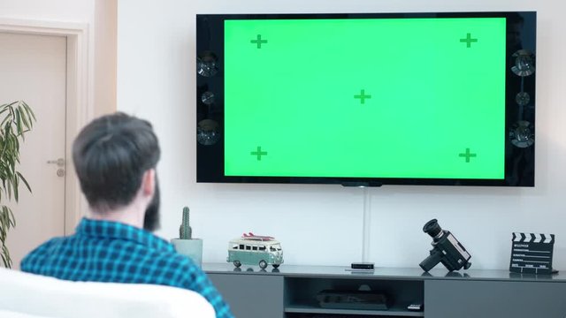 Man with a beard sits down on a couch relaxing and switchs on TV with green screen shot in 4k