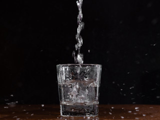 A glass of water on a black background, which is filled with a jet of water and flying spray