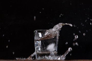 Naklejka na ściany i meble A glass of water on a black background, into which pieces of ice are thrown and splashes fly