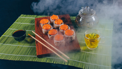 Delicious rolls with spicy sauce on wooden board with soy sauce and green tea on decorative bamboo napkin. Close up of tasty sushi rolls with chopsticks on cutting board with steam on black background