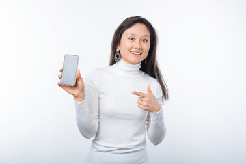 Photo of young happy woman pointing at smartphone while standing over white background