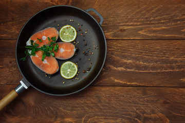 Cut pieces of raw red fish salmon in a cast iron pan with lemon slices spices and green parsley sprigs on a brown wooden background top view with an empty space for text, copy space, flat lay