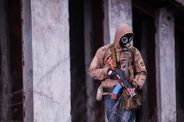 Armed man in gas mask, khaki jacket and blue jeans standing by the wall of destroyed building