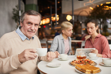 Portrait of mature man drinking tea and looking at camera while having lunch with his family in cafe