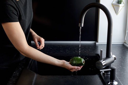 Woman Washes A Avocado Under Water In A Black Kitchen