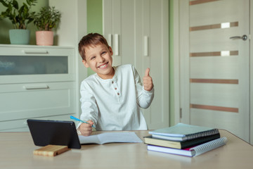 a boy with dark hair in a white shirt sits at a table and does homework using a tablet. distance learning at home, doing homework during the epidemic and pandemic covid-19