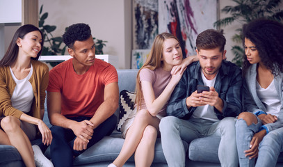Looking at the photos. Group of young happy multicultural people in casual wear looking at smartphone, enjoying time together while sitting on the sofa in the living room
