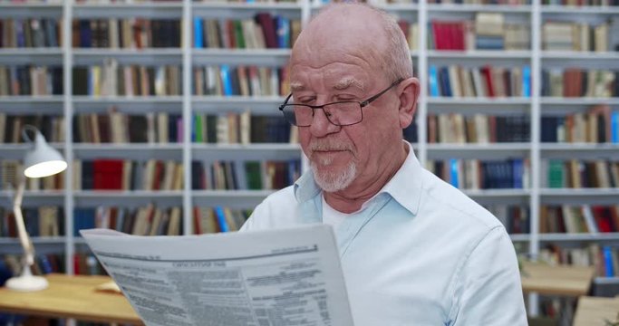 Old Caucasian bald man in glasses reading newspaper in library and smiling. Close up of male professor with gazette journal searching information in bibliotheca. Grandfather investigatin news.