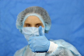 attractive female doctor in medical coat, in a cap and mask. Thumb lifted upwards, shows like on a blue background