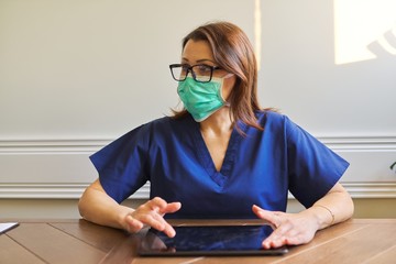 Fototapeta na wymiar Portrait of doctor woman in medical protective mask with digital tablet sitting at desk in office