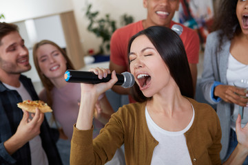Karaoke party. Beautiful young asian girl holding microphone and singing while playing karaoke with best friends at home, group of people having fun together, drinking and eating. Selective focus