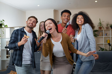 Young asian girl holding microphone and singing while playing karaoke with best friends at home. Group of people having fun together, drinking and eating pizza. Karaoke party