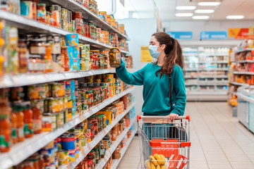 A young pretty Caucasian woman in a medical mask buys a preserved glass jar with vegetable mix at a...