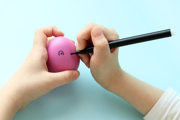 Caucasian child decorates an Easter pink egg. A child draws a cheerful face on a pink egg with a black marker. Selective focus.