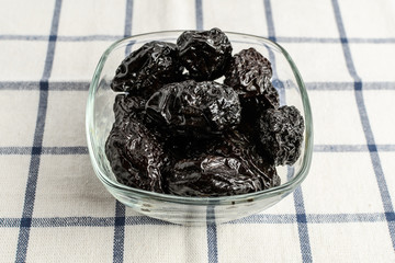 Dried plums in a transparent bowl on a table covered with white blue striped textile material, side view of healthy vegan food
