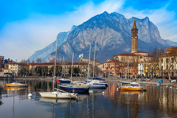 Fototapeta premium Lecco, Italy, February 16, 2020. The picturesque embankment of Lake Como in cloudy weather and the mountains behind it. Boats are reflected in the water