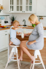 A little girl with mom, a young girl with a pregnant blonde, is poured and drink milk in the kitchen at the table