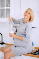 A young pregnant blonde in a gray dress sits on the kitchen table and fills a glass with milk from a jug