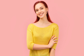 Nice attractive girl holds one hand on other arm by elbow, bows head, and laughs. Young girl with long red hair dressed in yellow sweater stands isolated on pink background in Studio with copy space