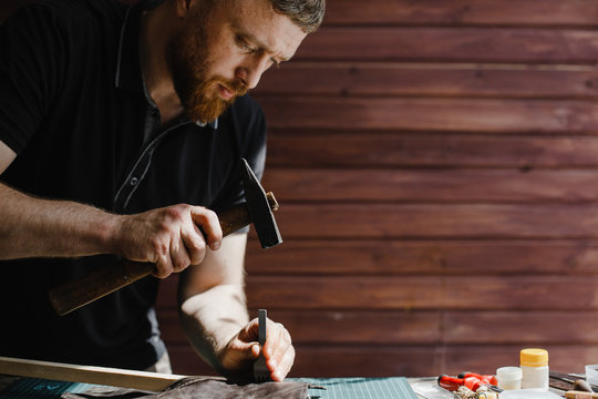 Craftsman with beard works in his workshop in daylight, using a hammer.