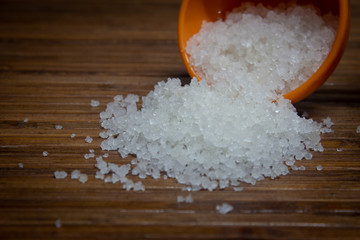 View of the rock salt crystals falling from a measuring cup over a brown wooden background