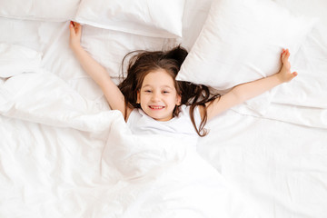 Happy little girl lies in bed at home in the morning. healthy baby sleep. white bed linen, space for text