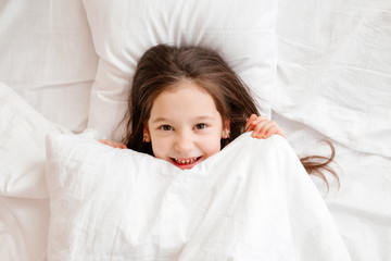 Obraz na płótnie Canvas Happy little girl lies in bed at home in the morning. healthy baby sleep. white bed linen, space for text