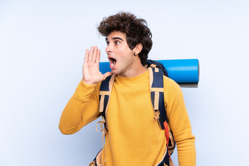 Young mountaineer man with a big backpack over isolated blue background shouting with mouth wide open