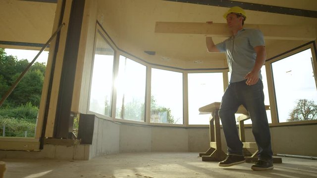 SLOW MOTION, LOW ANGLE, LENS FLARE: Cinematic shot of a young contractor carrying a plank across a modern CLT house under construction on a sunny day. Builder carries a wooden panel across sunlit room