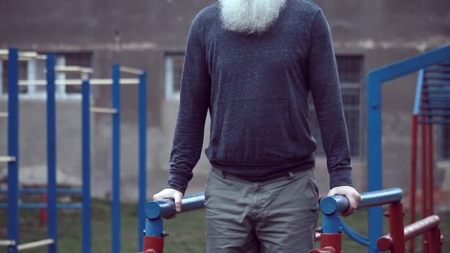 Senior bearded man with strong muscular arms exercising outdoors, doing pull ups, an elderly male performing pulling exercise on horizontal bar