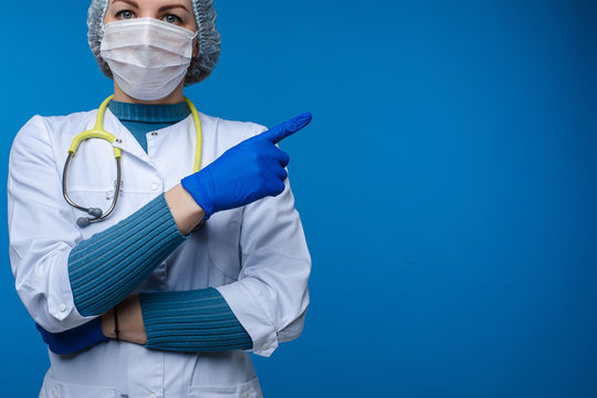 Stock photo portrait of a professional doctor or lab worker in PPE - personal protection equipment - mask, coat, gloves and hat. Doctor pointing at copy space on blue background.
