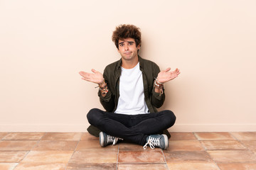 Young caucasian man sitting on the floor having doubts while raising hands